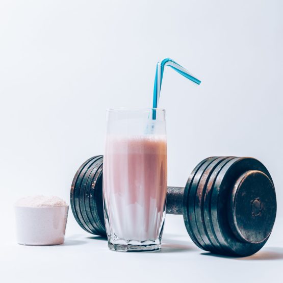 Glass of protein shake with a straw next to a portion of dry powder in a scoop and dumbbell. Sports drink and heavy weights.